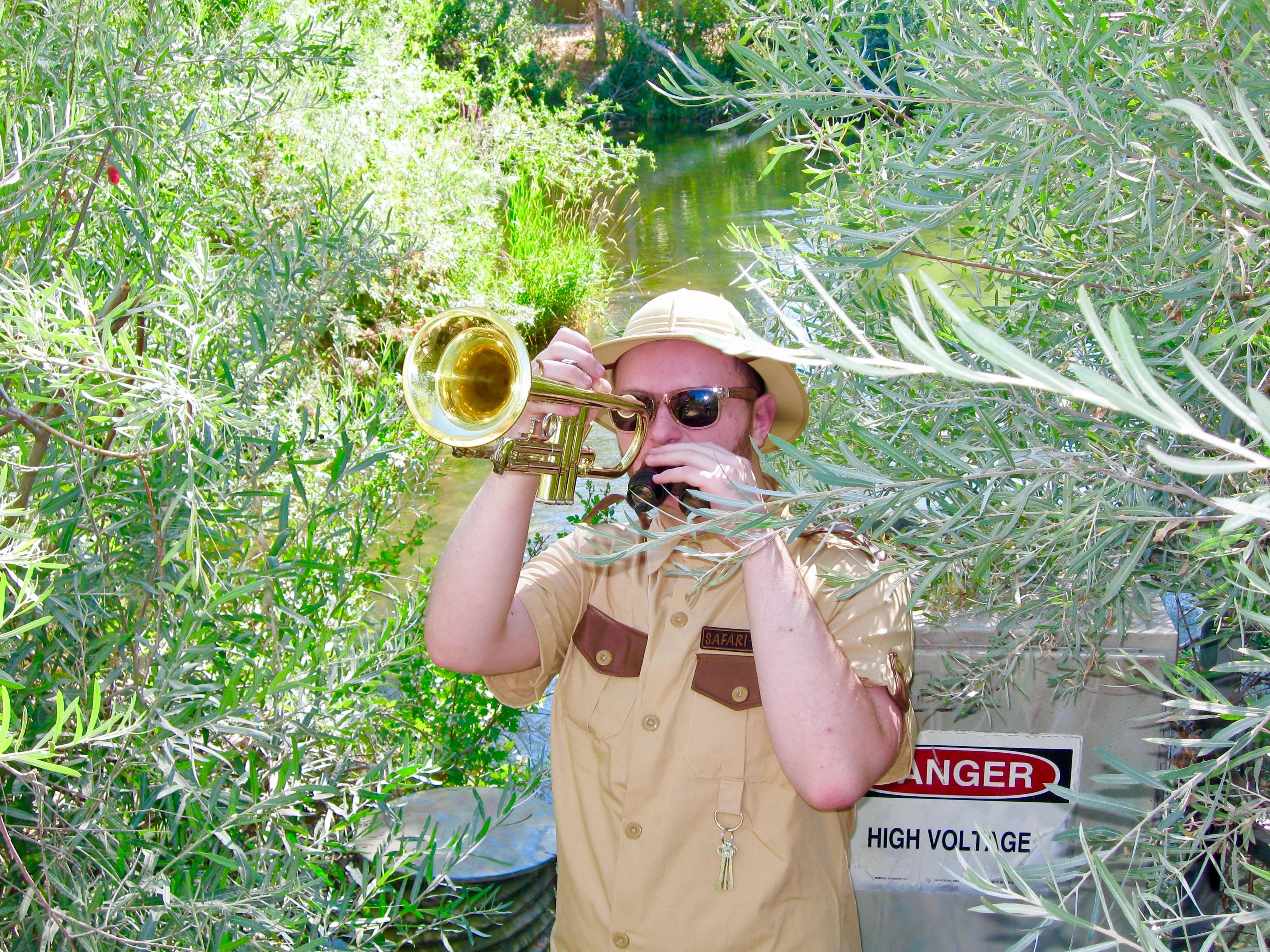 Daughter Lotus, wearing a safari uniform and sunglasses, playing a trumpet towards the camera.