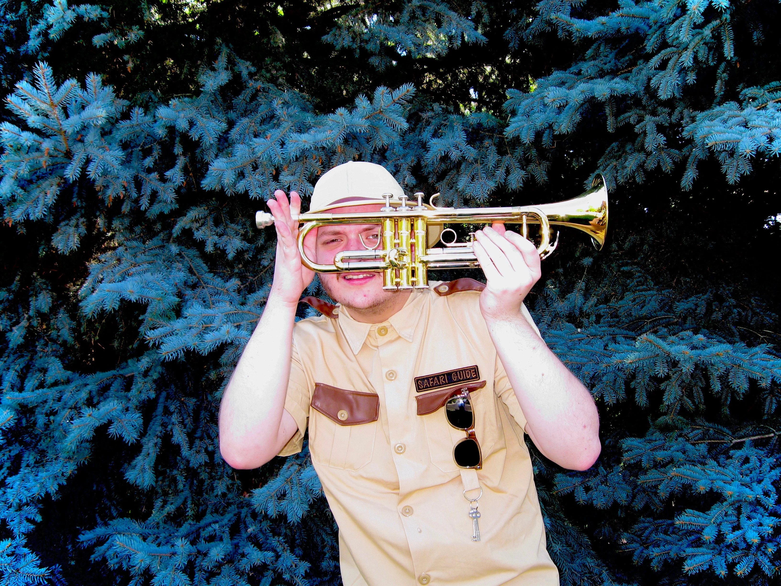 Daughter Lotus, wearing a safari uniform, looking through a gap in a trumpet while holding it in front of him with both hands. He's also standing in front of dark-blue fir trees.