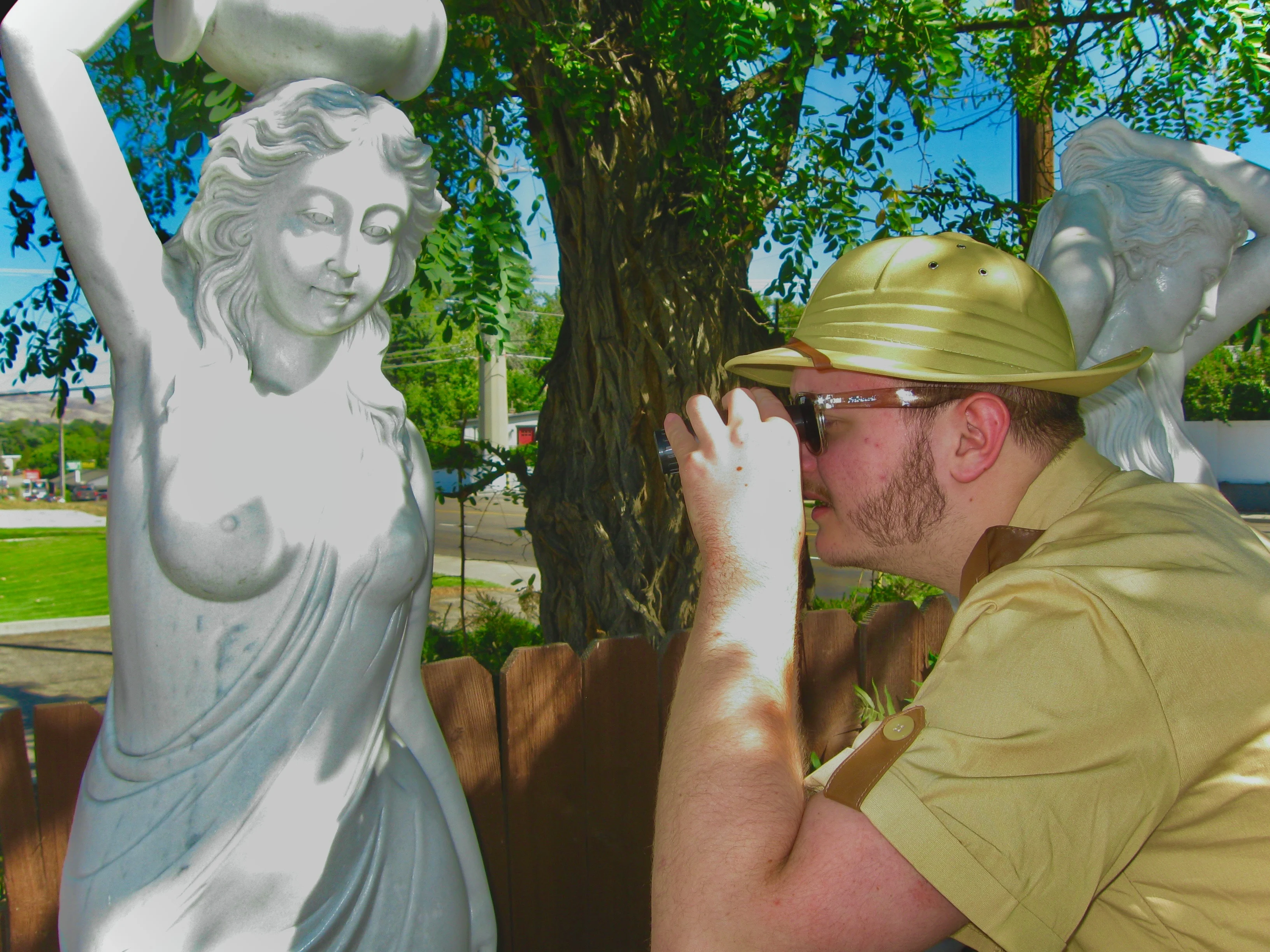 Daughter Lotus, wearing sunglasses and a safari uniform, looking through binoculars at a statue's bare breasts that are directly in front of him.