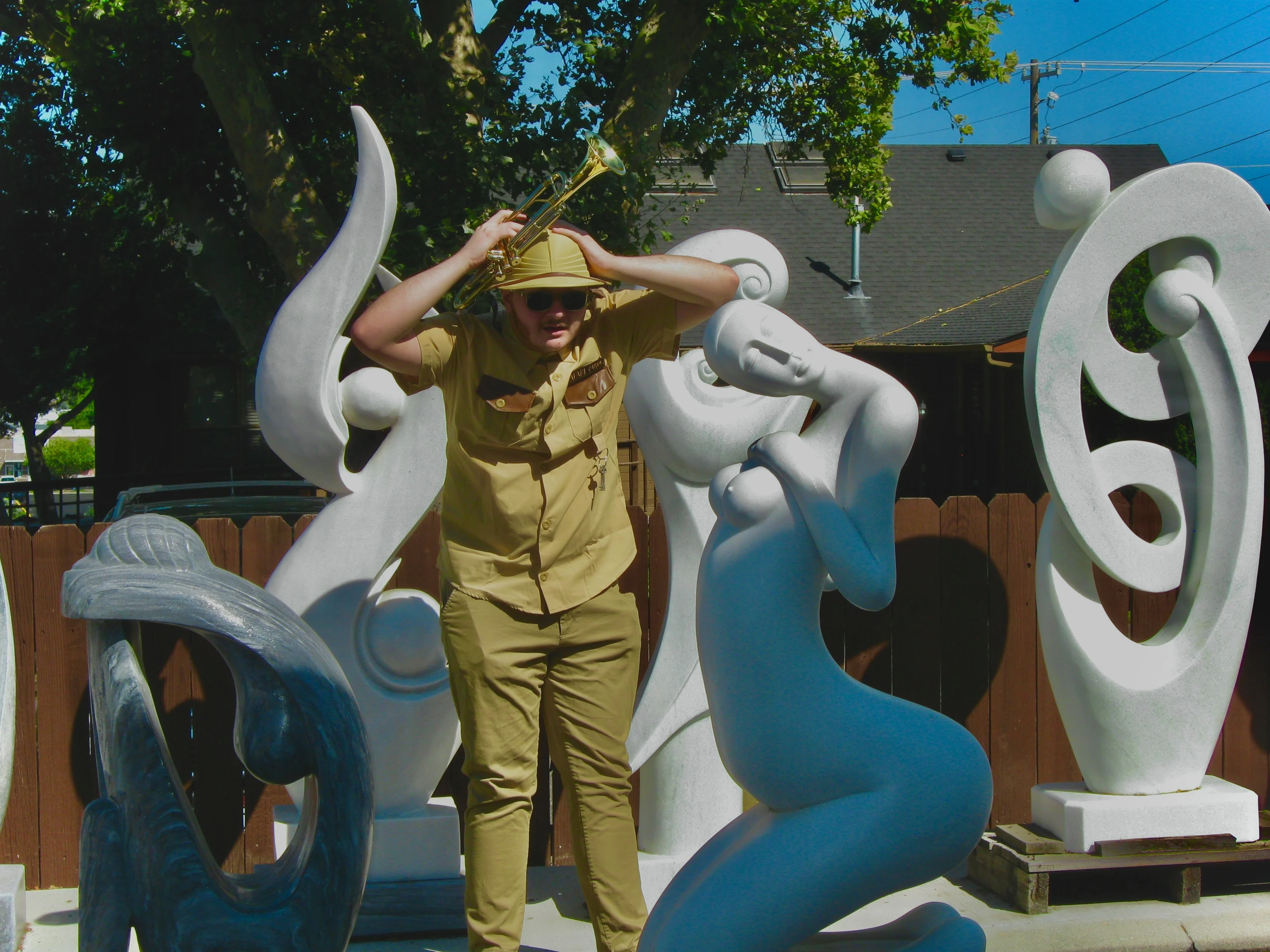 Daughter Lotus wearing sunglasses and a safari uniform, putting his hands on his head with a trumpet in his right hand, looking towards the camera with sunglasses as if he just witnessed something shocking.