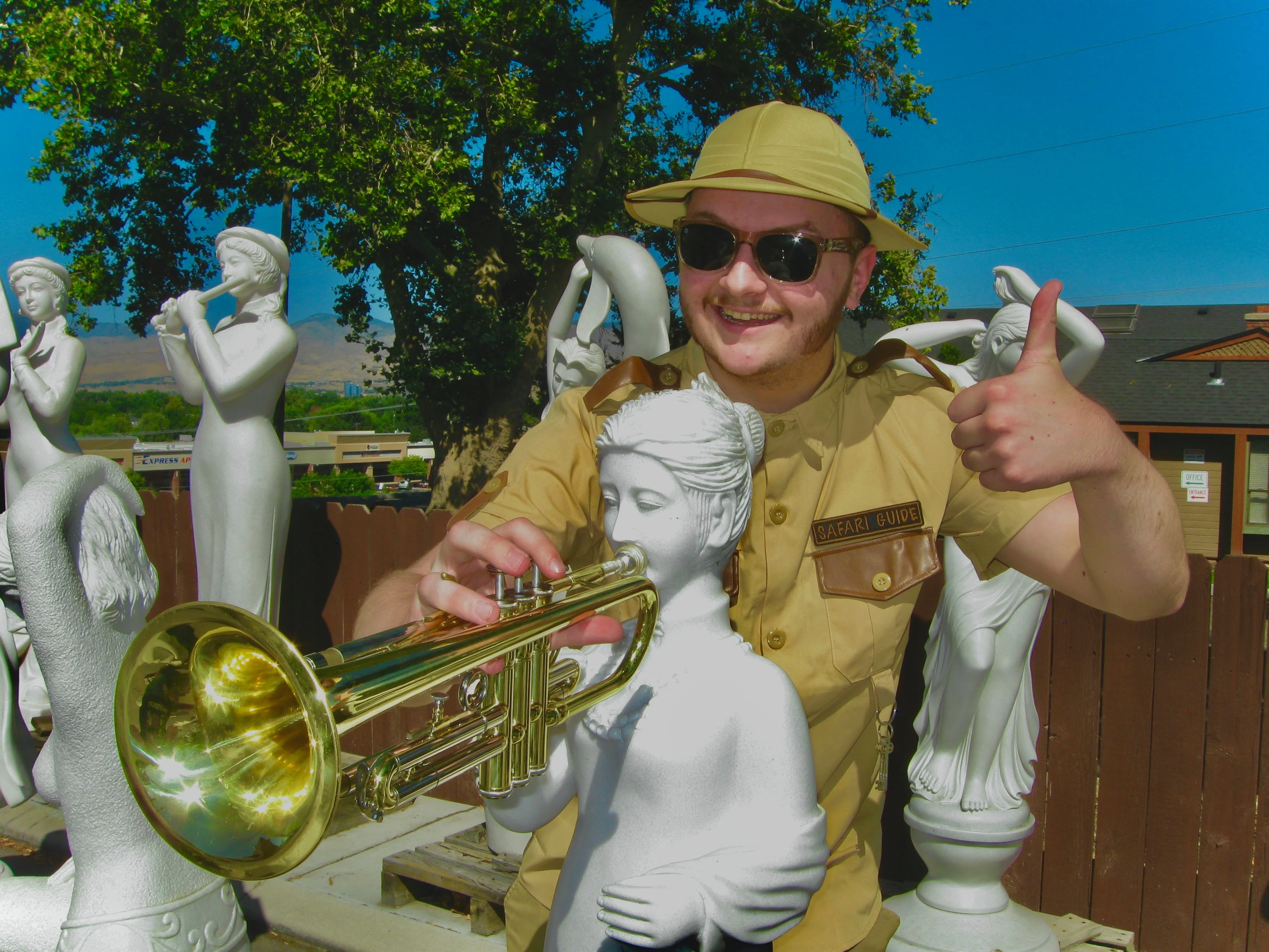 Daughter Lotus giving a thumbs-up while wearing sunglasses and a safari uniform, placing a trumpet in front of a statue to make it look as if the statue is playing it.